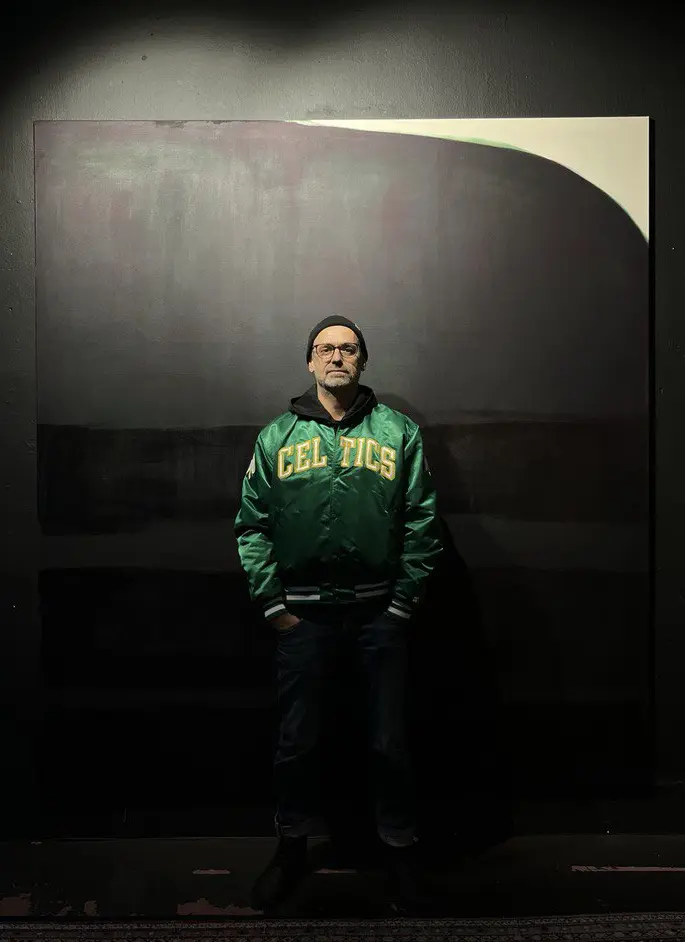 Jörg Mächtel steht in grüner Boston Celtics Jacke in einer Galerie vor einem großflächigen Bild.